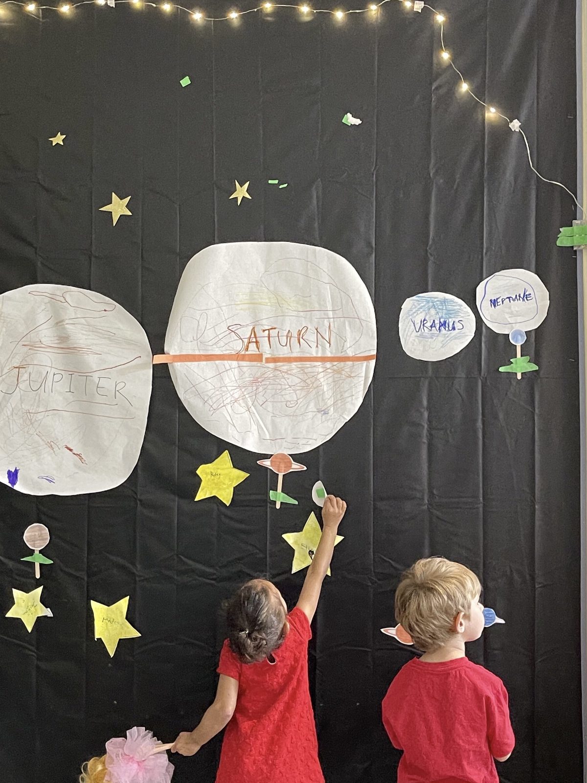Two children adding paper stars to a classroom space-project wall of hand-drawn planets